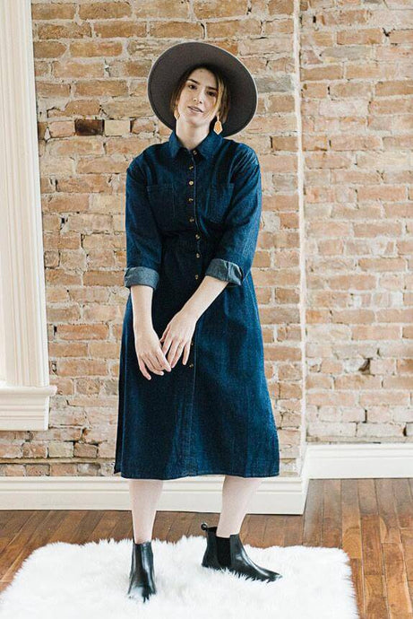 Woman wearing midi length dark denim dress with long sleeves that are rolled and button front closer detail front.