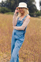 woman smiling standing in a grass field wearing wide leg baggy denim overalls 90s style.
