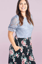 Woman wearing plus size modest style maxi dress with light blue lace bodice and floral pattern skirt with ruffled hem with her hands in dress pockets side.
