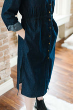 Woman wearing midi length dark denim dress with long sleeves that are rolled and button front closer detail with hands in dress pockets.