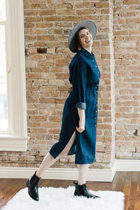 Woman wearing midi length dark denim dress with long sleeves that are rolled and button front closer detail and slits on side.