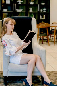Woman sitting reading a book wearing white mini dress with embroidered floral flare bell sleeves side.